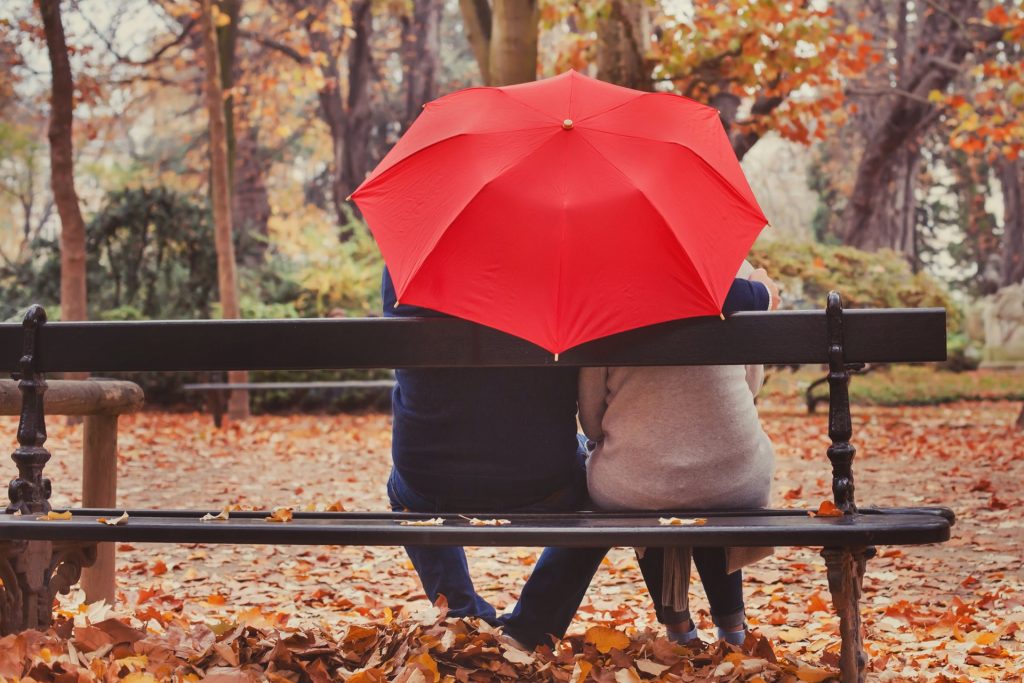Two people sitting on a park bench holding a red umbrella